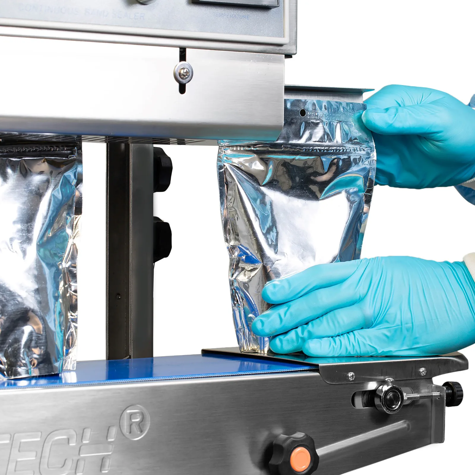un operador con guantes azules insertando una bolsa plástica plateada en la máquina selladora continua de banda JORES TECHNOLOGIES® para sellarla cerrada