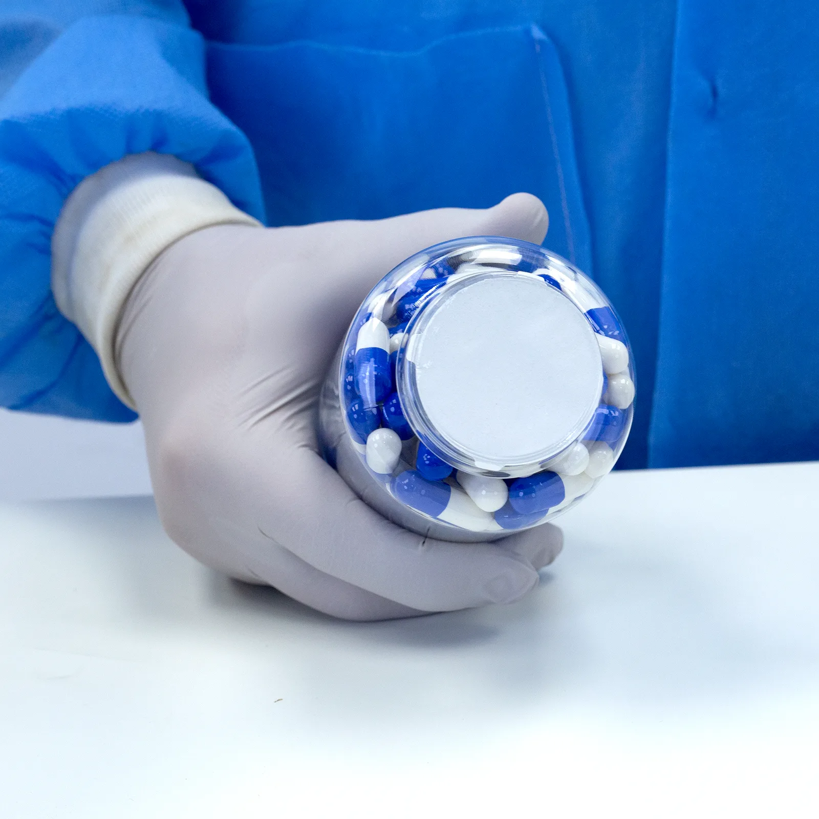 Persona que lleva guantes de nitrilo y sostiene un envase cerrado con un liner de sellado por inducción con pestañas. El envase está lleno de tabletas medicinales azules y blancas