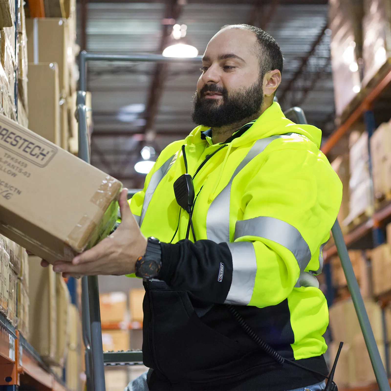 Un trabajador que lleva un suéter de seguridad amarillo de alta visibilidad clase 2 tipo R. Está retirando una caja de un contenedor en un entorno de almacén.